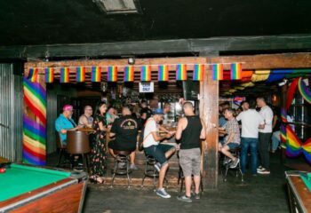 Mineshaft Bar scene with gay men, rainbow flags, pool tables, and a cash-only bar. Gay travel destination.