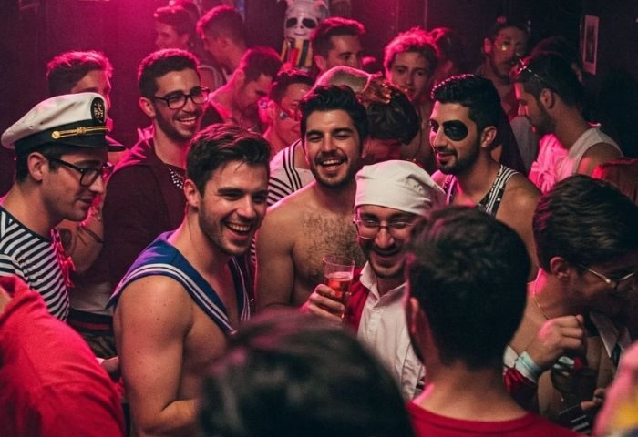 Crowd at Moeem Barcelona, a gay bar in Barcelona. Men in sailor costumes and casual wear smile and socialize under pink lighting.
