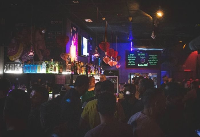 Moeem gay bar in Barcelona. A crowded bar with people standing near the counter, drinks, and a cocktail menu. Nightlife scene.