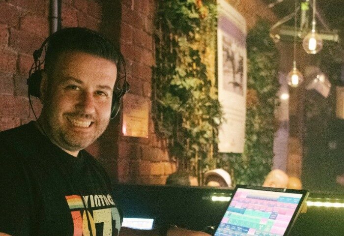 DJ at Mollie's Bar and Diner, Melbourne. Gay nightlife scene. Man smiling, wearing headphones, with laptop. Brick wall and greenery backdrop.