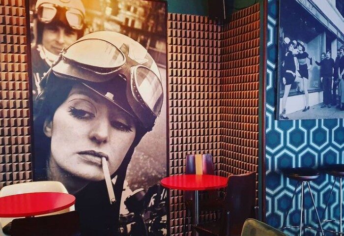 Mono Bar Milan interior. Retro design, woman with helmet poster, acoustic foam wall, and geometric wallpaper. Gay travel.