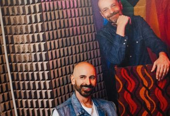 Mono Bar Milan portrait. Two smiling men, one in a denim vest, the other in a shirt, pose in front of a geometric wall. Gay travel.