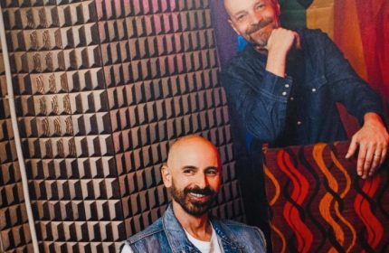 Mono Bar Milan portrait. Two smiling men, one in a denim vest, the other in a shirt, pose in front of a geometric wall. Gay travel.