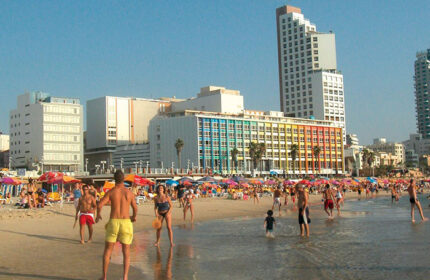 Dan Tel Aviv Hotel: Beachfront scene with people enjoying the water and sand. Luxury hotel in the background. Gay-friendly travel.