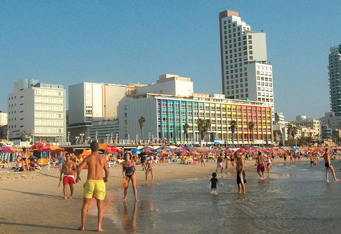 Dan Tel Aviv Hotel: Beachfront scene with people enjoying the water and sand. Luxury hotel in the background. Gay-friendly travel.