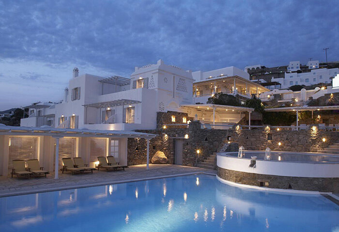 Porto Mykonos Hotel pool at dusk. Most-booked gay honeymoon hotel in Mykonos Town city center, Greece. White buildings, stone walls, lights.