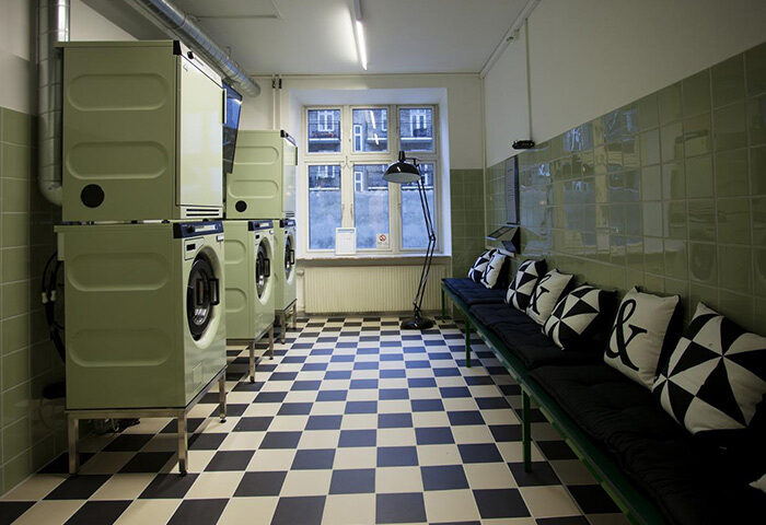 Urban House Copenhagen by MEININGER's laundry room. Washing machines, tiled floor, bench with pillows. Gay hostel with washing machine.