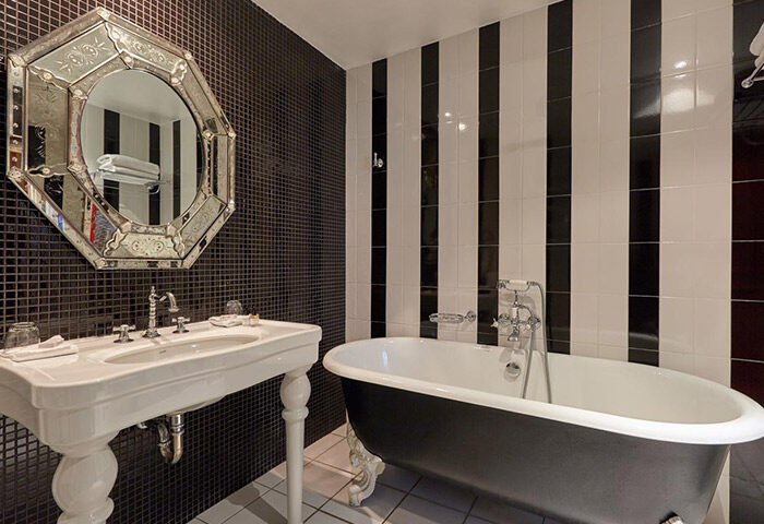 Hotel du Petit Moulin bathroom. Black & white striped tiles, black mosaic wall, vintage mirror, & clawfoot tub. Most-booked gay hotel in Paris.