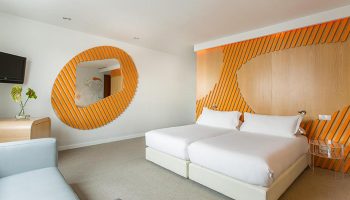 Room Mate Oscar hotel bedroom in Madrid's Chueca gayborhood. Modern design with twin beds, orange accents, and unique mirror. Top gay hotel.