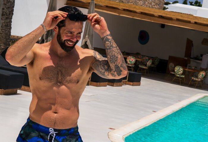 Akkadian Gay Only Hotel in Mykonos: Man smiling, shirtless with tattoos near pool. One of the most booked gay hotels in Mykonos.