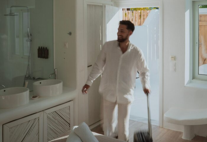 Akkadian Gay Only Hotel Mykonos: Man with luggage in all-white suite. Most-booked gay hotels in Mykonos beach offer luxury and comfort.