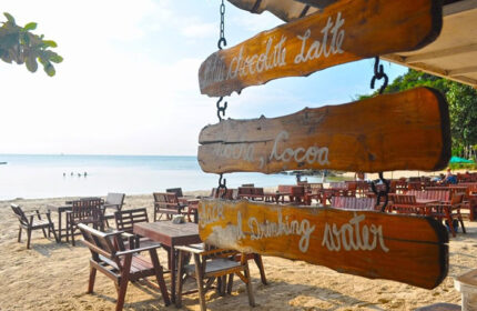Tubtim Resort beachfront cafe, Koh Samet. Wooden signs advertise drinks. Tables & chairs on the sand. Popular gay nightlife nearby.