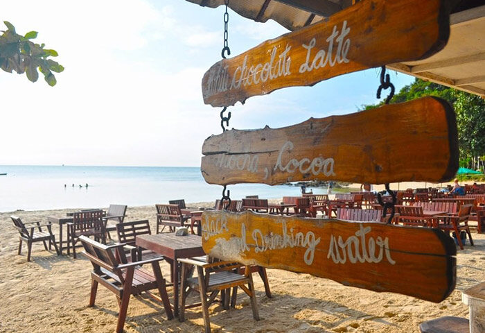 Tubtim Resort beachfront cafe, Koh Samet. Wooden signs advertise drinks. Tables & chairs on the sand. Popular gay nightlife nearby.