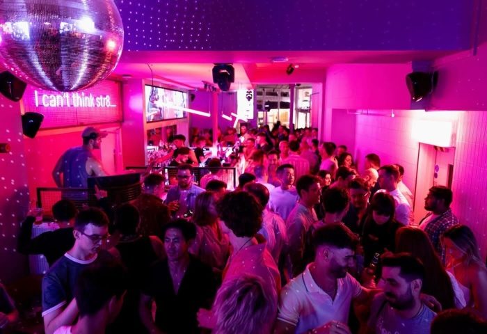 Crowded KU Bar / KU Klub, a popular gay bar in London. Disco ball, pink lighting, and a DJ create a vibrant atmosphere. Nightlife scene.