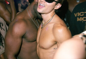 Gay New York: Men dancing shirtless at The Rosemont, a popular gay bar in NYC. A man in sunglasses smiles at the camera. Nightlife scene.
