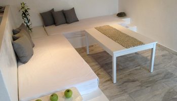 M for Mykonos gay hotel room near windmills. White interior with built-in seating, cushions, table, and apples. Gay travel advice.