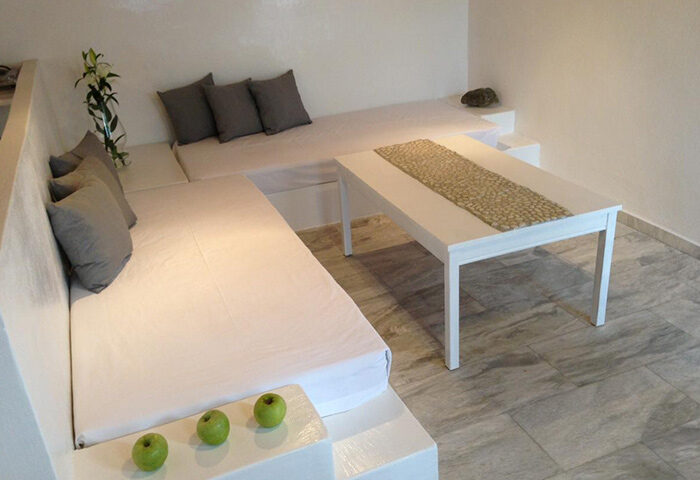 M for Mykonos gay hotel room near windmills. White interior with built-in seating, cushions, table, and apples. Gay travel advice.