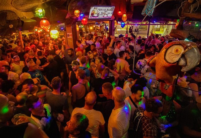Crowded Mr. Flamingo gay bar in Puerto Vallarta. Many men are standing, with colorful lights and a parrot statue visible.