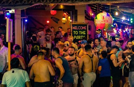 Mr. Flamingo Gay Bar in Puerto Vallarta. Crowded scene with shirtless men, Welcome to the Party sign, and festive lights. Gay travel.