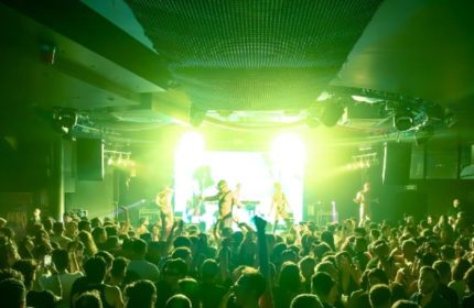 Muccassassina Rome: A vibrant gay club scene with a crowd enjoying the show. Green lights illuminate the stage and dance floor.