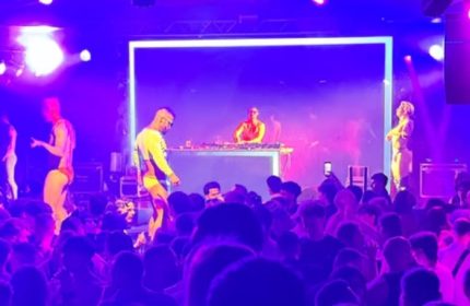 Muccassassina Rome: DJ and dancers perform on stage at a crowded gay party. Purple lighting and energetic atmosphere. Gay travel in Rome.