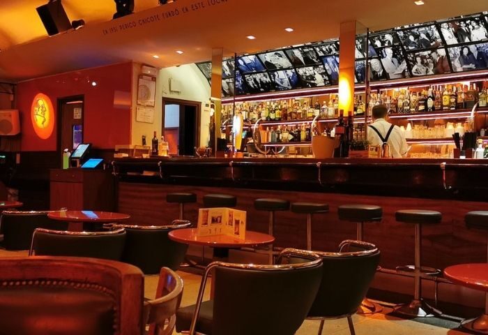 Interior of Museo Chicote, a gay bar in Madrid. Bar stools line the counter, with tables and chairs in front. Shelves of liquor behind the bar.