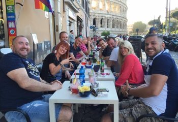 Gay travel: Group of friends at a bar in Rome, Italy, with the Colosseum in the background. Rainbow flag visible. My Bar Rome.