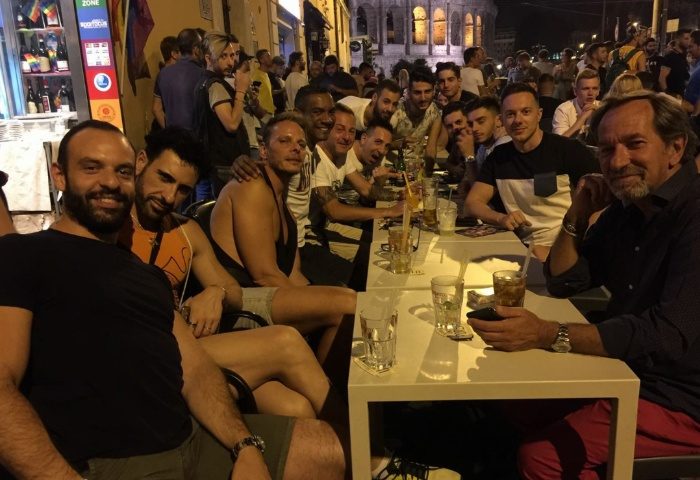 Gay men at a bar in Rome, Italy. Group of friends enjoying drinks at a table outside. Nightlife scene with the Colosseum in the background.