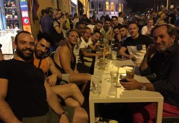 Gay men at a bar in Rome, Italy. Group of friends enjoying drinks at a table outside. Nightlife scene with the Colosseum in the background.