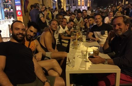 Gay men at a bar in Rome, Italy. Group of friends enjoying drinks at a table outside. Nightlife scene with the Colosseum in the background.