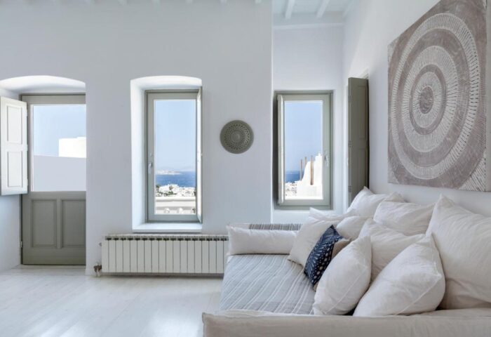 Myconian Inn gay budget hotel room. White interior with sea view, windows, sofa, pillows, and art. Mykonos travel for gay men.