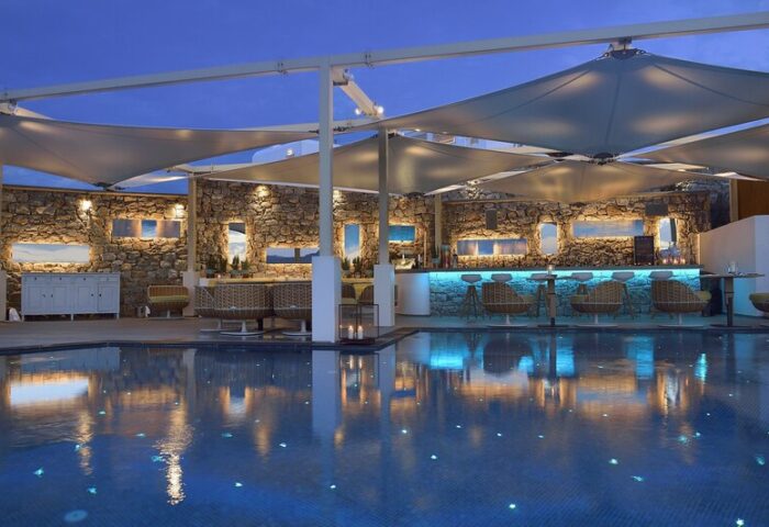 Myconian Korali, Mykonos gay hotel. Poolside bar at dusk with stone wall, blue lighting, and reflections in the pool. Gay travel.