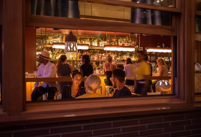 New York bar scene: View through window of people at a bar. Bottles line the shelves, lights hang above. Gay travel advice.