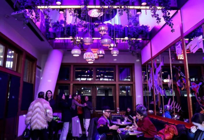 Interior of a New York bar with purple lighting, hanging plants, and stylish lamps. People are socializing at tables. Gay travel advice.