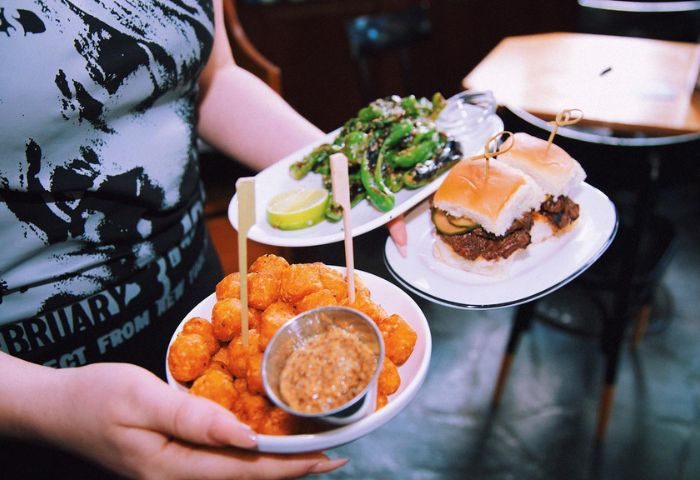 Waiter holding plates of food at NO BAR in New York. Tater tots, sliders, and shishito peppers. Gay travel food.