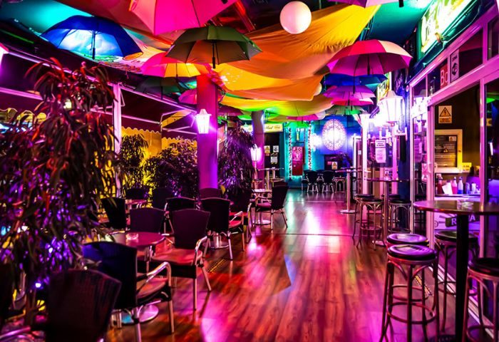 Interior of Na Und Bar, a gay bar in Gran Canaria. Colorful umbrellas hang from the ceiling. Tables and chairs are arranged on the dance floor.