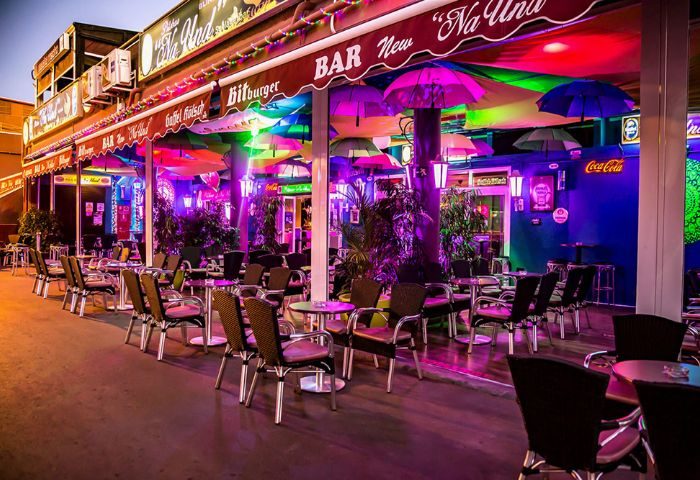 Na Und Bar, a gay bar in Gran Canaria. Outdoor seating area with colorful umbrellas, tables, and chairs. Nightlife for gay men travel.
