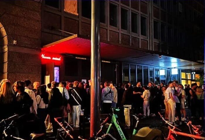 Night view of Never Mind Bar, Copenhagen. Crowd of gay men outside. Bicycles parked nearby. Red neon sign. Gay travel advice.