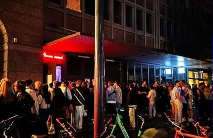 Night view of Never Mind Bar, Copenhagen. Crowd of gay men outside. Bicycles parked nearby. Red neon sign. Gay travel advice.