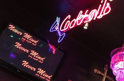 Never Mind Bar, Copenhagen. Neon 'Cocktails' sign, TV screen displaying the bar's name, and a chandelier in this gay night club.