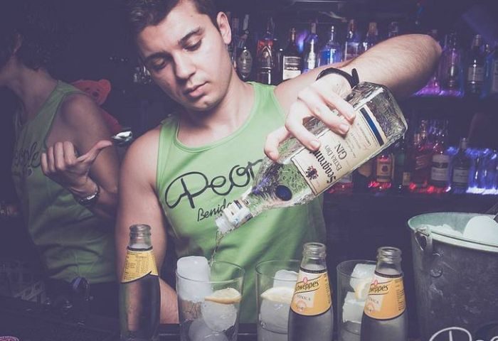 Bartender pours gin at New People gay bar in Benidorm, Spain. Person wears green tank top. Bottles of tonic and ice visible. Nightlife.