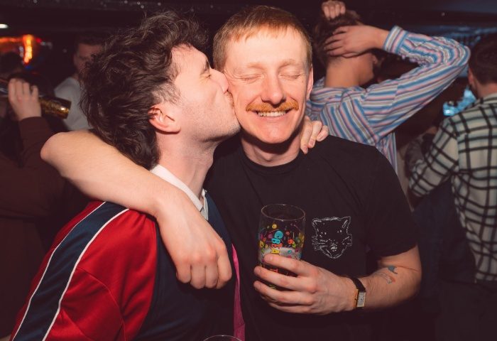 Gay couple in New York New York, Manchester. Man kisses partner's cheek. Nightlife scene, travel advice. LGBTQ+ friendly bar. Fun, love, joy.