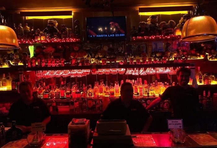 Nicho Bears Bar gay party in Mexico. Bartenders serve drinks at a bar lined with bottles and teddy bears under red lighting.