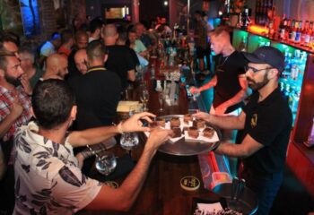 Night Barcelona Gay Cruise Club: Busy bar scene with men, bartender serving desserts, vibrant nightlife, gay travel destination.