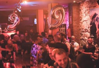 Night Barcelona Gay Cruise Club: Interior view of a crowded gay bar in Barcelona, Spain. Men gather at the bar, illuminated by red lighting.