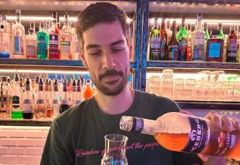 Gay bartender pours drink at NoLoSo Bar, Milan. Colorful bottles line shelves. Gay travel advice.