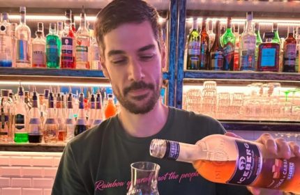 Gay bartender pours drink at NoLoSo Bar, Milan. Colorful bottles line shelves. Gay travel advice.