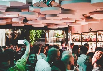 Crowded NoLoSo Bar in Milan. Gay bar scene with people, unique ceiling design, and bar shelves. Gay travel advice.