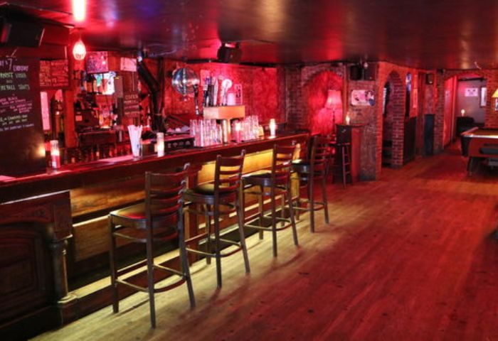 Interior shot of Nowhere Bar in New York. A gay bar with a long bar, stools, red lighting, and a pool table in the back.