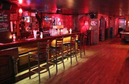 Interior shot of Nowhere Bar in New York. A gay bar with a long bar, stools, red lighting, and a pool table in the back.